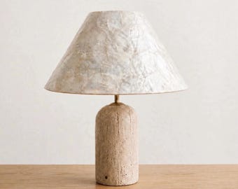 Può includere: Una lampada da tavolo con base cilindrica in pietra beige porosa e testurizzata. La lampada presenta un paralume conico composto da pezzi di conchiglia bianca iridescente sovrapposti, collegato alla base da un sottile stelo in ottone lucido.