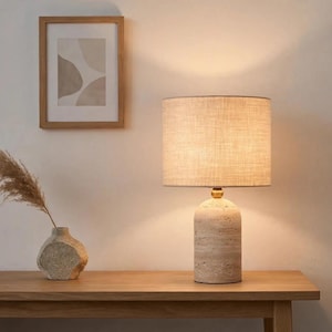 Pu&ograve; includere: Una lampada da tavolo con base cilindrica in pietra travertino, un piccolo dettaglio in metallo dorato e un paralume a tamburo in tessuto beige. La lampada emette una luce calda che illumina l'area circostante con una luce soffusa e neutra.