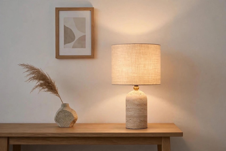 Pu&ograve; includere: Una lampada da tavolo con base cilindrica in pietra travertino, un piccolo dettaglio in metallo dorato e un paralume a tamburo in tessuto beige. La lampada emette una luce calda che illumina l'area circostante con una luce soffusa e neutra.