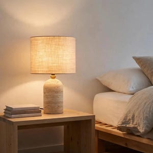 Pu&ograve; includere: Una lampada da tavolo con una base cilindrica in pietra beige porosa dalla finitura naturale e testurizzata, completata da un collo in metallo dorato. La lampada presenta un paralume cilindrico in tessuto beige chiaro che emana luce.