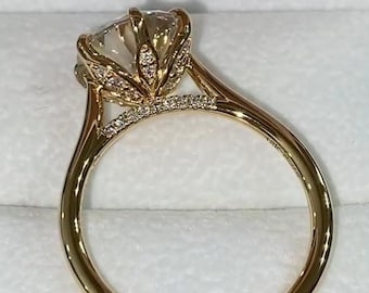 Puede incluir: Un anillo de oro con una gran piedra central redonda y transparente sujeta por un engaste en forma de pétalo. El engaste y la parte superior de la banda están incrustados con pequeñas piedras blancas brillantes.