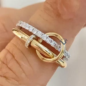 Puede incluir: Un anillo bicolor que presenta una banda de color dorado pulido entrelazada con una segunda banda incrustada con piedras transparentes y brillantes. El diseño incorpora un elemento de bucle circular que conecta ambas bandas.