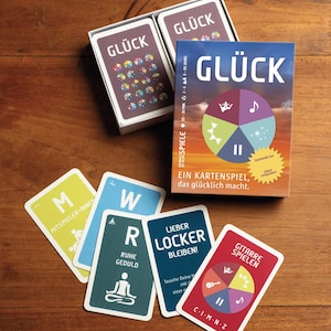 Könnte beinhalten: Ein Kartenspiel namens GLÜCK mit einer Schachtel, die ein buntes Rad-Grafikdesign zeigt, Text wie EIN KARTENSPIEL, DAS GLÜCKLICH MACHT, sowie mehrere einzelne Spielkarten mit Buchstaben, Symbolen und deutschen Phrasen wie RUHE GEDULD und LIEBER LOCKER BLEIBEN.