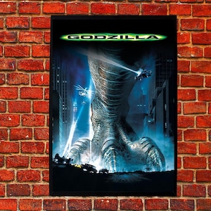 Può includere: Un poster cinematografico incorniciato del film "Godzilla" contro un muro di mattoni rossi. Il poster presenta una grande immagine dettagliata del piede e della parte inferiore della gamba di Godzilla, con un paesaggio urbano ed elicotteri sullo sfondo. Il titolo "GODZILLA" è in alto.