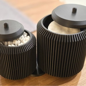 Può includere: Due contenitori cilindrici neri con coperchi, uno aperto e uno parzialmente. I contenitori hanno un design a coste. Uno contiene cotton fioc bianchi, l'altro un dischetto di cotone. I contenitori sono su una superficie di legno.
