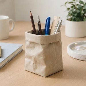 Puede incluir: Un portalápices de papel beige con textura arrugada, lleno de bolígrafos, lápices y una regla. Una taza blanca, un cuaderno y una pequeña planta en maceta también están sobre la superficie de madera.