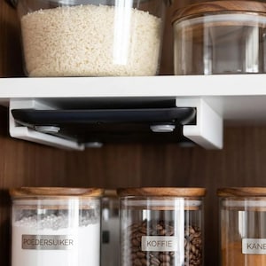 Pode incluir: Uma balança de cozinha digital preta montada sob uma prateleira branca usando um suporte de plástico branco. A balança possui uma superfície plana com pés circulares visíveis, posicionada entre potes de vidro com arroz, café e especiarias.