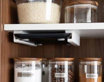 May include: A black digital kitchen scale mounted underneath a white shelf using a white plastic bracket. The scale features a flat surface with visible circular feet, positioned between glass storage jars containing rice, coffee, and spices.
