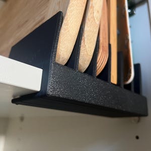 Op de afbeelding: Een zwarte, getextureerde kunststof organizer bevestigd aan een plank, voorzien van meerdere verticale sleuven waarin verschillende lichtgekleurde houten snijplanken rechtop staan voor opslag in een keukenkast.