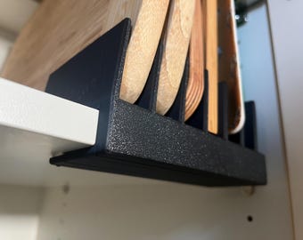 May include: A black, textured cutting board organiser installed inside a cabinet. The organiser holds several wooden cutting boards of various shapes and sizes. The organiser is mounted on a white shelf.