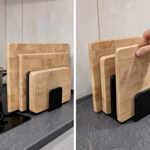 Pode incluir: Um conjunto de tábuas de corte de madeira de vários tamanhos, guardadas num suporte de metal preto. As tábuas são feitas de madeira clara e são empilhadas verticalmente. Uma panela de aço inoxidável está sobre uma placa de fogão preta.