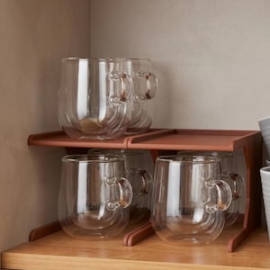 Op de afbeelding: Heldere glazen mokken met handvatten staan op een houten plank. De mokken hebben een dubbelwandig ontwerp. De plank is roodbruin en wordt ondersteund door een houten frame. De mokken zijn leeg.