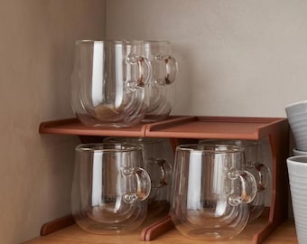 May include: Several clear glass mugs with handles are arranged on a wooden shelf. The mugs have a double-walled design. The shelf is reddish-brown and supported by a wooden frame. The mugs are empty, ready for use.