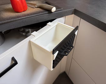 May include: A white plastic kitchen waste bin with a black front panel featuring a perforated grid pattern and a small handle. The bin is designed to hang over the top edge of a cabinet door, providing a compact storage solution for kitchen scraps.