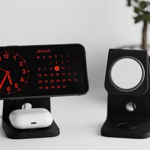Op de afbeelding: Zwarte oplaadstandaards voor een telefoon, oortelefoons en smartwatch. De telefoon toont een rode klok en kalender voor januari. De oortelefoons zijn wit. Een kleine plant in een zwarte pot staat op de achtergrond.