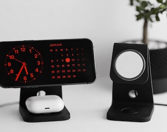 May include: Black charging stands for a phone, earbuds, and smartwatch. The phone displays a red clock and calendar for January. The earbuds are white. A small plant in a black pot is in the background.