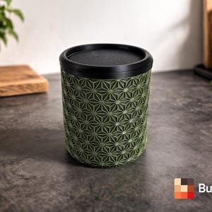 Può includere: Un contenitore cilindrico verde con un motivo geometrico, sormontato da un coperchio e un bordo neri. Il contenitore è su una superficie grigio scuro. Il logo "Builted3D" è visibile in basso a destra.