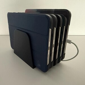 Op de afbeelding: Een zwarte oplaadstation houdt meerdere blauwe tablethoezen vast. Een grijze oplaadkabel is aangesloten op de tablets. Het station staat op een wit oppervlak.
