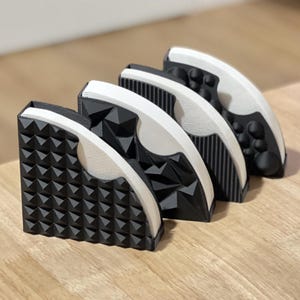 Può includere: Quattro porta filtri per caffè geometrici in bianco e nero sono disposti su una superficie di legno. Ogni supporto presenta un motivo strutturato unico, tra cui piramidi, triangoli, linee verticali e protuberanze arrotondate. I supporti sono leggermente angolati.