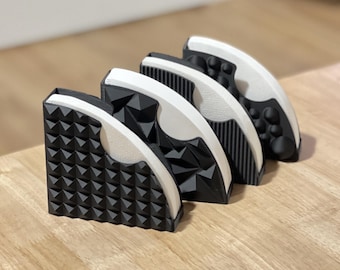 May include: Four black and white geometric coffee filter holders are arranged on a wooden surface. Each holder has a unique textured pattern, including pyramids, triangles, vertical lines, and rounded bumps. The holders are angled slightly.