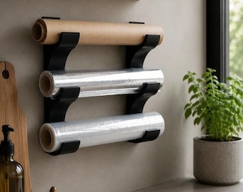 May include: A black metal kitchen wrap organiser holds three rolls of paper, foil, and cling film. The organiser is mounted on a light grey wall above a countertop. A wooden shelf and a potted plant are also visible.