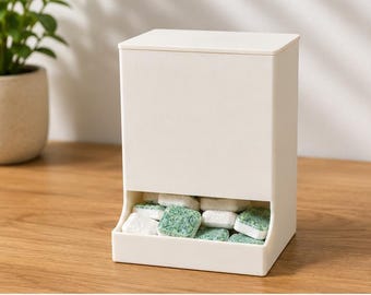 May include: A rectangular, off-white plastic storage container with a flat lid and an open bottom section. The lower compartment is filled with several small, square-shaped dishwasher detergent tablets that have a white and green speckled surface.