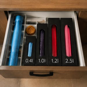 Pode incluir: Um organizador de gaveta de cozinha contendo um rolo azul, um objeto redondo amarelo texturizado e quatro dispensadores retangulares pretos rotulados 0.4l, 1.0l, 1.2l e 2.5l. Os dispensadores contêm sacos plásticos visíveis através de aberturas.