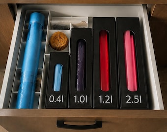 May include: A kitchen drawer organiser containing a blue roll, a round yellow textured object, and four black rectangular dispensers labelled 0.4l, 1.0l, 1.2l, and 2.5l. The dispensers hold blue, purple, red, and pink plastic bags visible through cutouts.