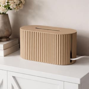 Op de afbeelding: Een beige kabelbox met een geribbeld ontwerp. De ovale doos heeft een sleuf aan de bovenkant en een gat aan de zijkant voor snoeren. Een witte kabel komt uit de zijkant. De doos staat op een wit oppervlak.