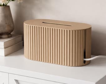 May include: A beige cable tidy with a ribbed design. The oval-shaped box has a slot on top and a hole on the side for cords. A white cable extends from the side. The box is on a white surface.