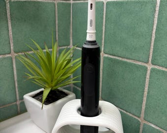 May include: A black electric toothbrush with a white brush head, in a white holder with wooden legs. A small potted plant is next to it. The background has green square tiles.