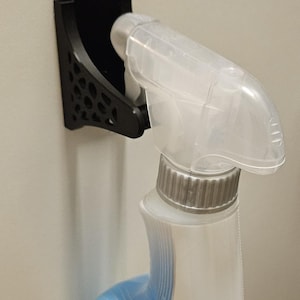 Könnte beinhalten: Ein schwarzer, 3D-gedruckter Sprühflaschenhalter, der zum Aufhängen an einer Tür oder einem Schrank konzipiert ist. Der Halter hält eine Sprühflasche sicher. Die Flasche ist weiß mit blauen Akzenten und einer transparenten Sprühdüse.