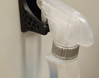 May include: A black, 3D-printed spray bottle holder designed to hang over a door or cabinet. The holder securely supports a spray bottle. The bottle is white with blue accents and a clear spray nozzle. The holder is made of plastic.