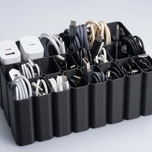 Può includere: Un organizer per cavi in plastica nera con più scomparti, riempito con vari cavi di ricarica e adattatori di alimentazione. I cavi sono di diversi colori e stili, tra cui bianco, nero e grigio. Due adattatori di alimentazione bianchi sono visibili in alto a sinistra.