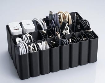 May include: A black cable tidy with numerous compartments, containing a variety of charging cables and power adapters. The cables are in diverse colours and styles, including white, black, and grey. Two white power adapters are visible in the top left corner.