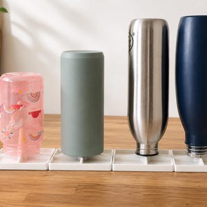Peut inclure: Quatre bouteilles d'eau de différentes couleurs et designs sont présentées sur une surface en bois. Les bouteilles comprennent une rose avec un motif arc-en-ciel, une grise, une en acier inoxydable et une bleu marine. Chaque bouteille est posée sur une base carrée blanche.