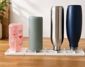 May include: Four water bottles in various colours and styles are arranged on a wooden surface. The bottles include a pink one with a rainbow pattern, a grey one, a stainless steel one, and a navy blue one. Each bottle is placed on a white square base.