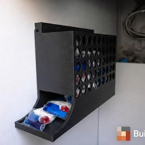 Op de afbeelding: Een zwarte, aan de muur gemonteerde dispenser voor wasmiddelcapsules. De dispenser heeft een honingraatpatroon aan de voorkant en een bakje aan de onderkant. Er zijn verschillende wasmiddelcapsules zichtbaar in het bakje. Het logo "Builded3D" staat rechtsonder.