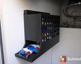 May include: A black, wall-mounted laundry detergent pod dispenser. The dispenser features a honeycomb design and a tray at the bottom. Several detergent pods are visible in the tray. The logo "Builded3D" is in the bottom right corner.