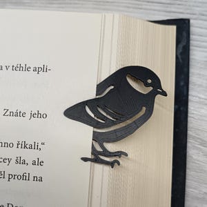 Op de afbeelding: Een zwart vogelvormig boekenlegger van een stevig materiaal, geplaatst op de rand van een boek. De boekenlegger is een silhouet van een vogel met gedetailleerde uitsparingen. De pagina's van het boek zijn lichtbeige.