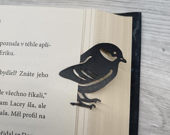May include: A black bird-shaped bookmark, made of a solid material, is placed on the edge of a book. The bookmark is a silhouette of a bird with detailed cutouts. The book's pages are light beige.