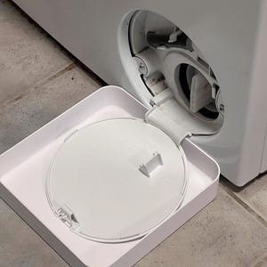 Peut inclure: Une machine à laver blanche avec la porte d'accès au filtre ouverte. Un bac blanc est positionné en dessous de l'ouverture, contenant le couvercle du filtre. La machine est sur un sol carrelé.