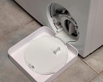 May include: A white washing machine with the filter access door open. A white tray is positioned below the opening, holding the filter cover. The machine is on a tiled floor.