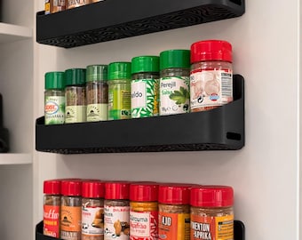 May include: Three black spice racks inside a white cupboard door, each holding several spice jars with colourful labels and lids. The jars contain various spices, including paprika, turmeric, and garlic.