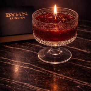May include: A lit red candle in a clear, ornate glass bowl with a stem. The candle is next to a dark book with gold lettering that reads "BYHN LUXURY". The candle is on a dark, marble-like surface.