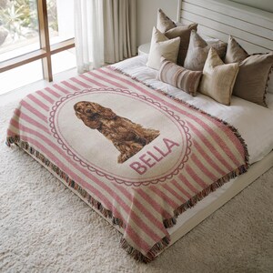 May include: A woven throw blanket featuring a pink and white striped pattern with a central oval frame containing an illustration of a brown spaniel dog and the name BELLA printed in pink capital letters, finished with a fringed edge.