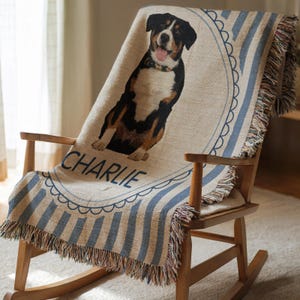 May include: A woven throw blanket featuring a dog portrait with the name "CHARLIE" in blue script. The blanket has blue and beige stripes and a fringed edge. The dog is black, brown, and white.