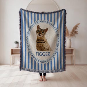 May include: A blue and white striped throw blanket featuring a portrait of a cat named Tigger. The cat is brown and tan with green eyes, set within an oval frame. The blanket has a fringed edge.