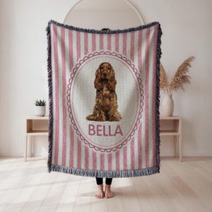 May include: A woven throw blanket with a pink and white striped pattern. The center features a brown Cocker Spaniel illustration inside a pink oval frame, with the name "BELLA" below. The blanket has fringed edges.