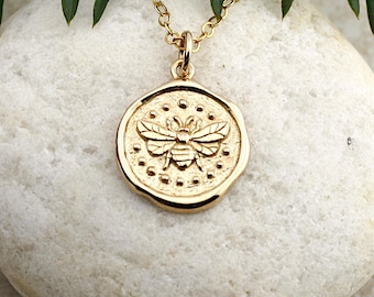 May include: A gold-colored pendant necklace featuring a circular charm with a detailed bee design. The bee is centered on the charm, surrounded by small dot details. The necklace is displayed on a white stone, with green foliage in the background.